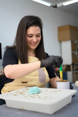 Stüdyoda mumlar yapan bir kadın. Etrafı aletler ve mumlarla çevrili. Eldiven ve önlük takıyor.