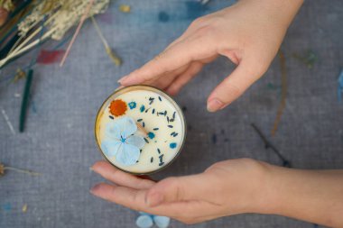 Sanatçının ellerinin yakın çekimi kurumuş çiçekler ve renkli bir zanaat atölyesinde diğer doğal elementlerle süslenmiş el yapımı bir mumu sunar.