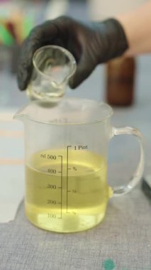 Eldiven giyen bir zanaatkar, küçük bir şişeden büyük bir şişeye sıvı balmumu döker, muhtemelen mum yapımına hazırlanır.