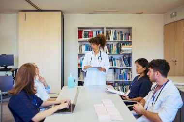 Doktorlar ve hemşireler aktif olarak tıbbi ekip toplantısına katılıyor, teknolojiden faydalanıyor ve modern bir hastane ortamında hasta bakımını tartışıyorlar.