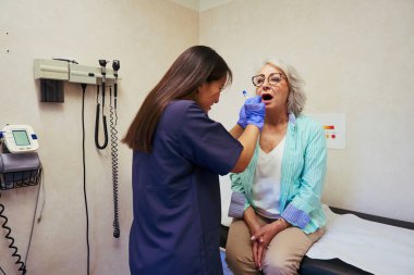 Eldiven giyen hemşire, klinikteki sağlık kontrolü sırasında yaşlı bir kadının boğazını incelemek için dil bastırıcı kullanıyor.