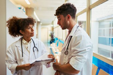 İki doktor hastane koridorunda hasta kayıtlarını tartışıyor. Sağlık hizmetlerinde takım çalışması ve işbirliği yapıyorlar.