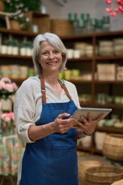 Çiçekçide çalışırken dijital tablet kullanarak siparişleri yöneten gülümseyen olgun bir kadın.
