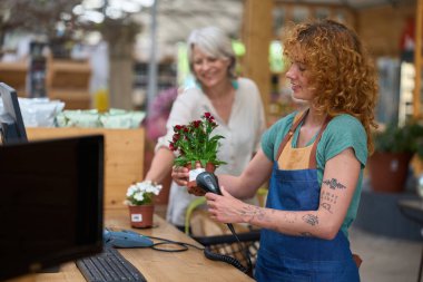 Çiçekçi, çiçeklerle dolu canlı bir çiçekçiden alırken müşterinin elinde tuttuğu saksı bitkisinin barkodunu tarıyor.