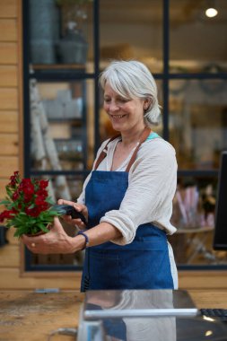 Kıdemli çiçekçi barkod tarayıcısıyla kırmızı çiçekleri tarıyor, çiçekçide çalışıyor, mavi önlük giyiyor.