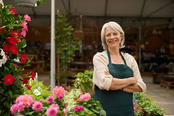 Kendine güvenen olgun bir çiçekçinin portresi çapraz kollarıyla poz veriyor, çiçeklerle çevrili bitki odasında.