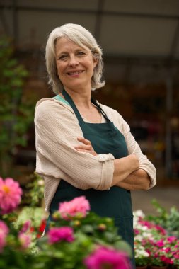 Renkli çiçeklerle çevrili çapraz kollarla poz veren kendinden emin bir çiçekçinin portresi, serada çalışıyor.