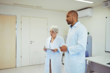 İki doktor, bir erkek ve bir kadın, bir hastane odasında, bir hastanın tıbbi tablosunu ve tedavi planını tartışıyorlar.