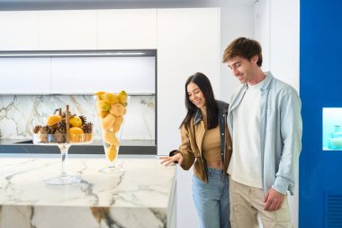 Modern mutfak galerisinde mermer tezgahı değerlendiren gülümseyen çift hayallerindeki evin içini planlıyorlar.