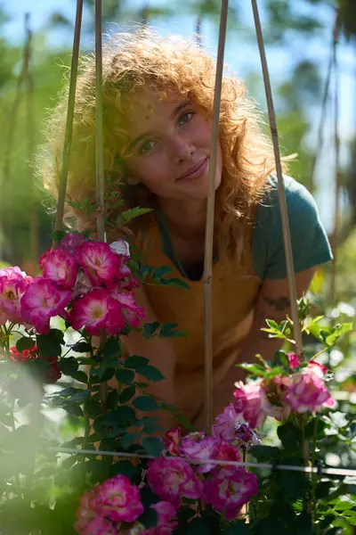 Güneşli bir bahçede, yemyeşil ve çiçek açan çiçeklerle çevrili canlı pembe güllere bakarken gülümseyen genç bir kadın.