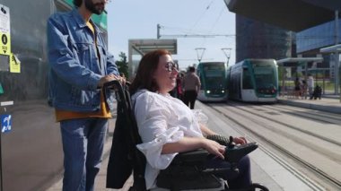 Çağdaş tramvay platformu yakınlarındaki gülümseyen engelli kadına tekerlekli sandalye yardımı sağlayan, kentsel hareketliliği somutlaştıran ve kapsayıcı ulaşım