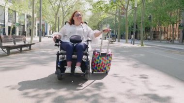 Elektrikli tekerlekli sandalyeyle şehir caddesinde gezen neşeli, iri yarı kadın, yaz tatili deneyimi sırasında valizini çekiyor ve bağımsızlık yayıyor.