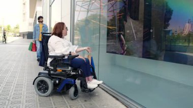 Özürlü bir kadın şehir kaldırımında güvenle geziniyor, destekçi bir arkadaşıyla vitrinlere bakıyor, tekerlekli sandalye kullanıyor, kapsayıcı yaşam tarzı ve toplumsal etkileşimi temsil ediyor.