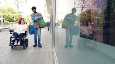 Sevgili çift birlikte vitrinlere bakmaktan hoşlanıyor, kadın tekerlekli sandalyeyle şehir merkezinde gezinirken partnerin birden fazla alışveriş çantası taşıması, ortak hareket kabiliyeti ve bağımsızlığı göstermesi.