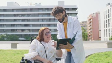 Dostane doktor, parkta tekerlekli sandalyede gülümseyen engelli bir kadına tabletle tıbbi bilgi veriyor.