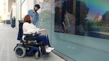 Elektrikli sandalyeyle şehir kaldırımında gezinen kendinden emin, güpegündüz vitrinlere bakarken destekleyici arkadaşıyla neşeli bir anı paylaşan iri yarı bir kadın.