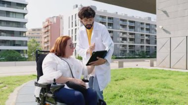 Tıbbi uzman tekerlekli sandalyedeki bir hastaya belgeleri gösteriyor, dışarıda sağlık durumunu tartışıyorlar.