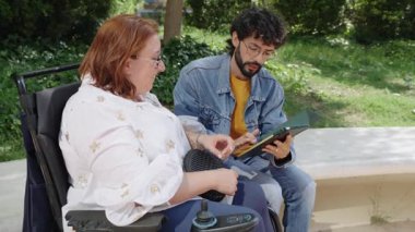 Tekerlekli sandalye kullanan bir kadın ve arkadaşı parkta bir bankta oturmuş tablete bakıyor ve konuşuyorlar.