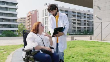 Doktor, dışarıda parkta tekerlekli sandalyedeki gülümseyen bir kadına tabletle tıbbi bilgileri açıklıyor.
