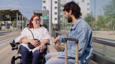 Tekerlekli sandalyedeki mutlu engelli kadın istasyonda toplu taşıma için beklerken bavul taşıyan turistle konuşuyor.