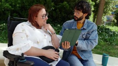 Tekerlekli sandalye kullanan bir kadın ve parktaki bankta oturan bir adam birlikte tablete bakıyor ve konuşuyorlar.