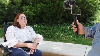 Kadın gazeteci neşeli bir kadınla röportaj yapıyor. Tekerlekli sandalye kullanıyor. Sunny Park 'ta kişisel öyküsünü paylaşıyor.