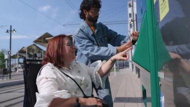 Nazik bir arkadaş, şehir tramvay istasyonunda bilet alırken tekerlekli sandalyedeki genç bayana yardım ediyor, güneşli bir günde ulaşılabilirliğini ve beraberliğini gösteriyor.