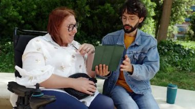 Tekerlekli sandalyedeki neşeli kadın tablet kullanıyor, park yeşilliği arasındaki arkadaşıyla anı paylaşıyor, arkadaşlığı ve teknolojiyi somutlaştırıyor.