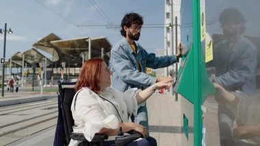 Elektrikli sandalyedeki genç kadın, tramvay bileti alırken arkadaşından yardım alırken, kentsel ulaşım istasyonunda ulaşılabilirliği ve bağımsızlığı vurguluyor
