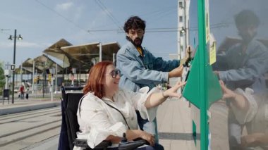 İstasyonda tekerlekli sandalyeden tramvay bileti alan neşeli bir kadın, şehir seyahati sırasında arkadaşından destek alıyor.