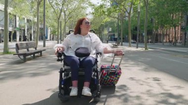 Elektrikli sandalyeyle şehir manzarasında gezen, bavulla gezen ve yaz tatili keşfi sırasında etrafa neşe saçan bir kadın gezgin.