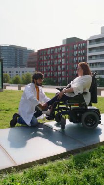 Tıbbi uzman, motorlu tekerlekli sandalyedeki bir kadına şehir parkında rahat edebilmesi için ayak sehpasını düzelterek yardım ediyor.