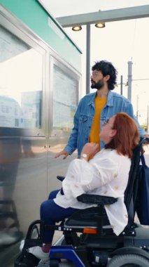 Tekerlekli sandalyedeki bir kadına yardım eden yardımsever bir adam istasyonda tren tarifesine bakıyor.