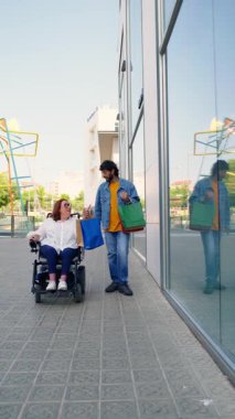Kız arkadaşına tekerlekli sandalyede yardım eden genç bir adam alışveriş çantalarını taşırken ve alışveriş merkezinin yanında yürürken