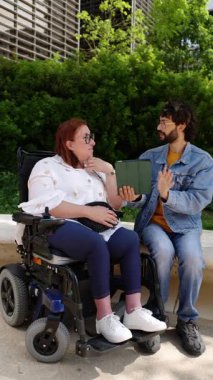 Tekerlekli sandalye kullanan neşeli bir kadın. Bir arkadaşıyla dijital tablet paylaşıyor. Birlikte dışarıda vakit geçiriyorlar.