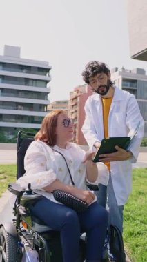 Dost canlısı doktor, parkta tekerlekli sandalyedeki mutlu engelli kadına tablet üzerinde tıbbi sonuçlar gösteriyor.