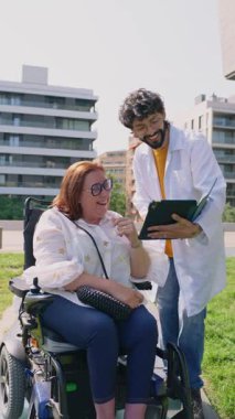 Tıbbi uzman, açık şehir ortamında tekerlekli sandalye kullanan gülümseyen bir kadınla iletişim kurarken tabletle ilgili sağlık bilgilerini paylaşıyor. Profesyonel destek ve hasta etkileşimine vurgu yapılıyor