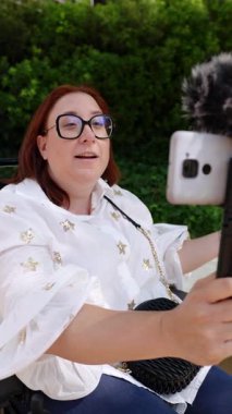 Kadın gazeteci dışarıda bir video röportajı kaydediyor, dış mikrofonu olan bir akıllı telefon kullanıyor ve selfie çubuğuna bağlı.