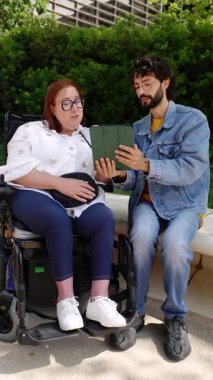 Tekerlekli sandalye kullanan bir kadın dışarıda arkadaşlarıyla vakit geçirmeyi, sohbet etmeyi ve tableti paylaşmayı sever.