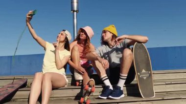 Neşeli genç arkadaşlar selfie çekerken, kaykay yaparken, kaykay yaparken ve kaykay yaparken okyanus arka planında güneşli mavi gökyüzünün altında, ahşap iskelede anıları yakalıyorlar.