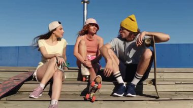 Üç neşeli arkadaş, iki kadın ve bir adam, tahta bir kaykay rampasında oturuyorlar, sohbet ediyorlar ve gülüyorlar, güneşli bir günün tadını çıkarıyorlar.