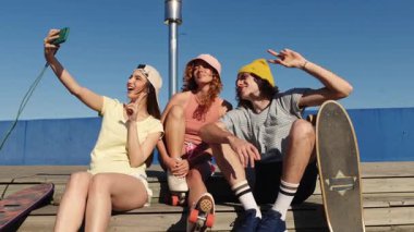 Neşeli bir grup arkadaş güneşli yaz gününde kaykay ve patenle iskelede selfie çekiyorlar.