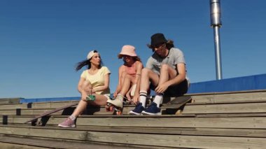 Genç arkadaşlar tahta tribünlerde otururken paten hazırlıyorlar açık mavi gökyüzü ve aktif yaşam tarzıyla güneşli bir yaz gününün tadını çıkarıyorlar.