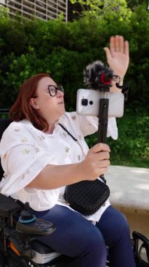 Profesyonel kızıl saçlı gazeteci canlı yayın yaparken uzaktan çalışıyor, yeşil park ortamı yakınlarındaki ulaşılabilir açık hava ortamından akıllı telefon ve dış mikrofon kullanıyor