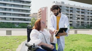 Tıbbi uzman, açık hava parkı konsültasyonu sırasında tekerlekli sandalyedeki gülümseyen kadın hastayla tabletle teşhis detaylarını tartışıyor.