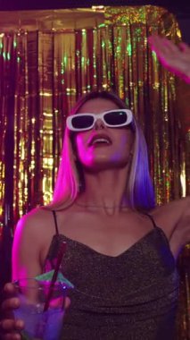 Elinde bir kokteyl ile dans eden genç bir kadın altın arka planda bir disko ortamında, yavaş çekim