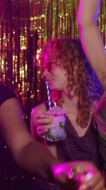 Kıvırcık kızıl saçlı ve kalp şeklinde güneş gözlüklü genç bir kadın disko partisinde kokteyl içiyor, ağır çekimde.