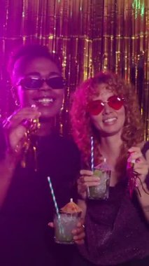 İki kadın gülümsüyor ve ağır çekimde altın folyolu bir partide kokteyl içiyorlar.