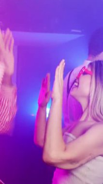 Değişik arkadaşlardan oluşan enerjik bir grup dans ediyor, alkışlıyor, renkli neon ışıklar altında neşe ve ritmik hareketlerle gece hayatını kutluyorlar.