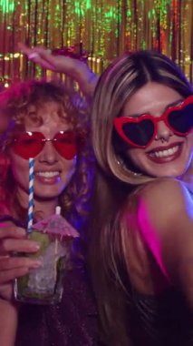 Kalp şeklinde güneş gözlüklü genç kadınlar renkli ışıklarla aydınlatılmış bir disko partisinde kokteyl içiyorlar.